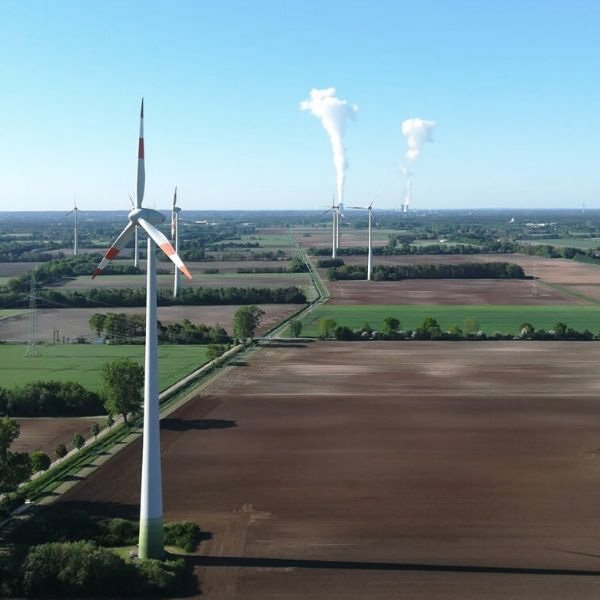 mehrere Windenergieanlagen stehen auf Feldern und Wiesen - im Hintergrund ist ein Kraftwerk zu sehen