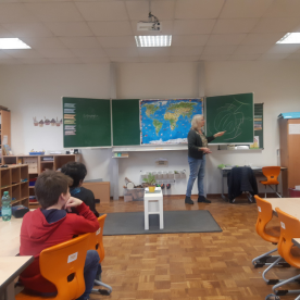 Lehrer erklärt an einer Tafel, Schüler sitzen in einem Klassenzimmer und hören zu.