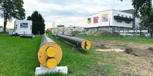 Große Rohre liegen auf einer Wiese, Baustelle und Gebäude im Hintergrund.