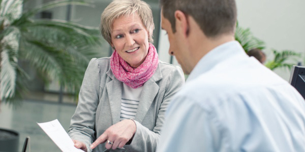 Eine Frau mit rosa Halstuch zeigt auf ein weißes Blatt Papier und schaut einen vor ihr stehenden Mann an.