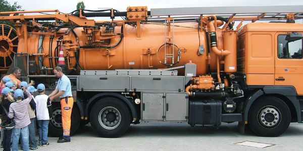 Ein Mann in orangener Warnkleidung und mehrere Kinder stehen vor einem organfarbenen LKW.