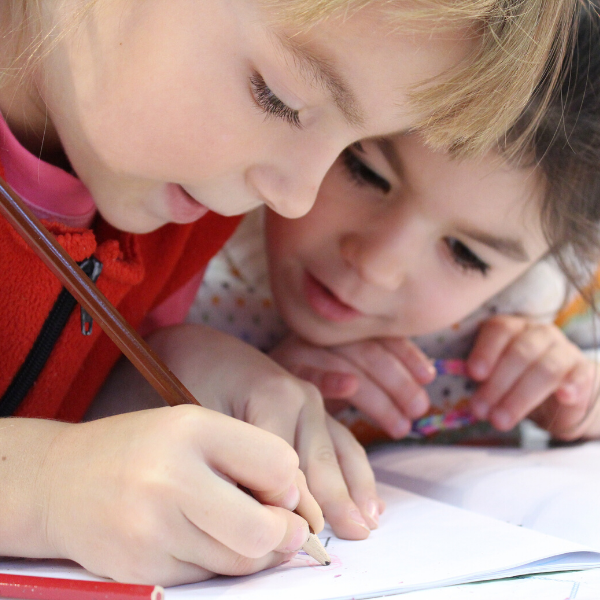 zwei Kinder schauen eng aneinandergepresst auf ein Blatt Papier, ein Kind schreibt mit einem länglichen Stift
