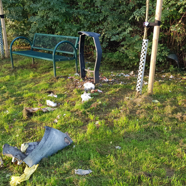 Zerbrochene Mülleimer und verstreuter Müll auf einer Wiese neben einer Bank.