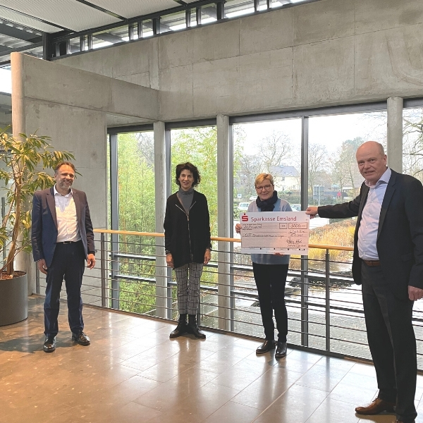 Vier Personen halten einen großen Scheck in einem modernen Gebäude.