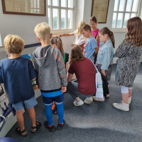 Gruppe von Kindern lauscht einer Erklärung in einem Raum mit großen Fenstern.