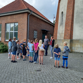 Kindergruppe steht draußen vor einem Backsteingebäude und hört einer Erklärung zu.